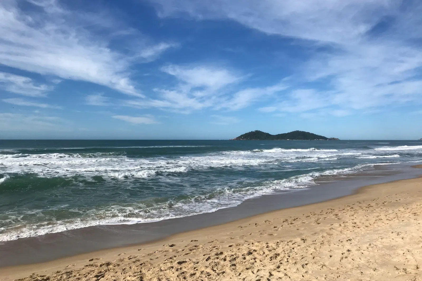 Praia do Campeche em Florianópolis, com ondas chegando à areia e a Ilha do Campeche ao fundo sob céu azul